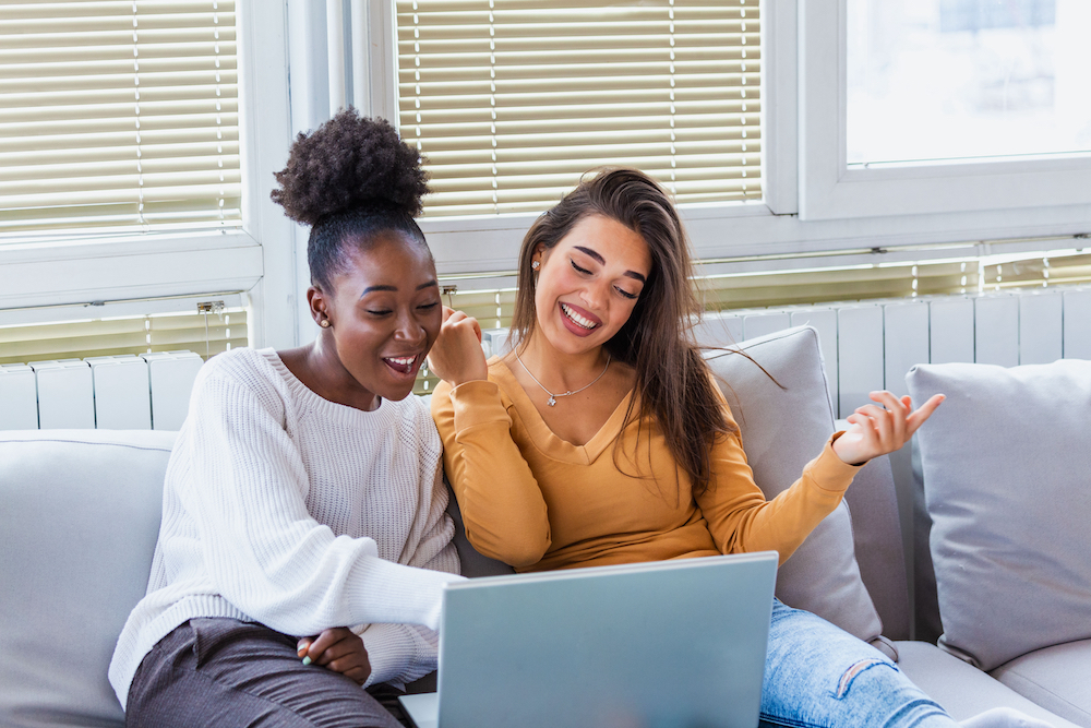 Two happy roommates laugh and smile while using a laptop